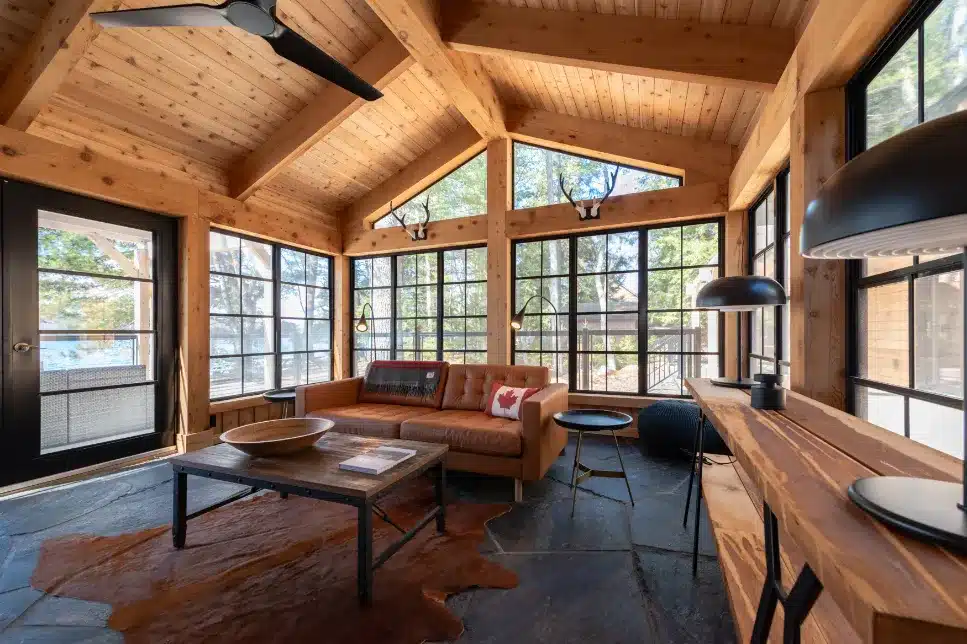 Spacious dark framed windows in a rustic Sunroom with wooden ceilings and a leather sofa, overlooking a forested view.