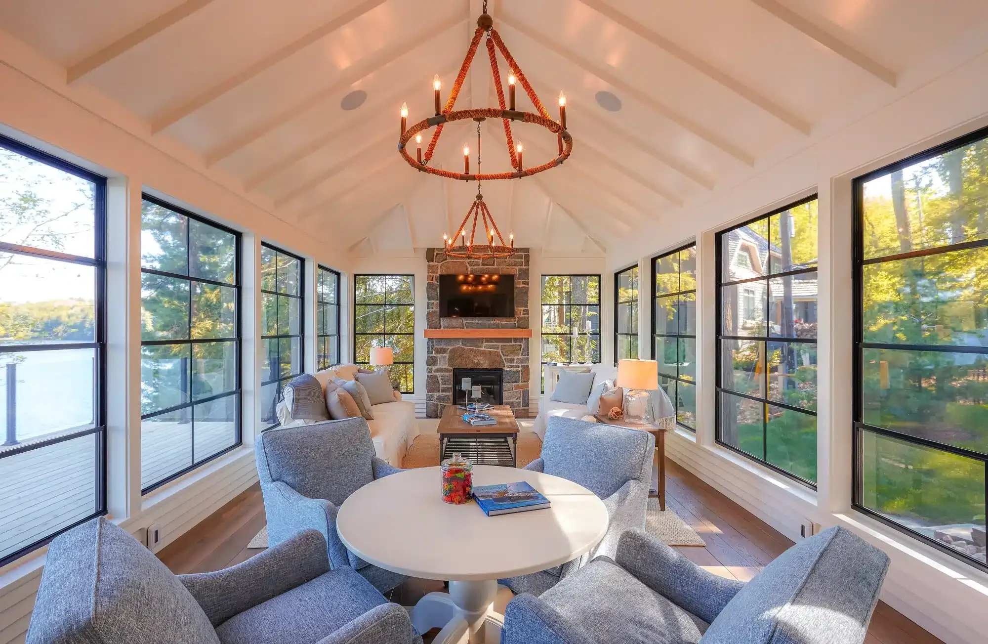 Sunroom with black-framed windows, a stone fireplace, and comfortable seating, showing beautiful lake views.