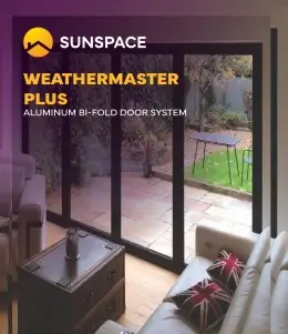 Sunspace WeatherMaster Plus aluminum bi-fold door system, black frame, opening onto a paved patio and green garden.