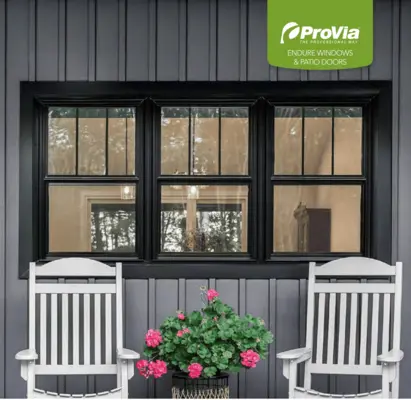 Three black-framed double-hung windows with top-only grilles set against dark grey vertical siding, with two white rocking chairs and pink flowers in the foreground.