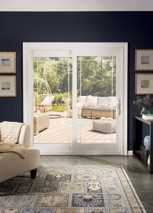 A white sliding glass door with a perimeter grid pattern, looking out from a dark navy room onto a sunlit deck with patio furniture.