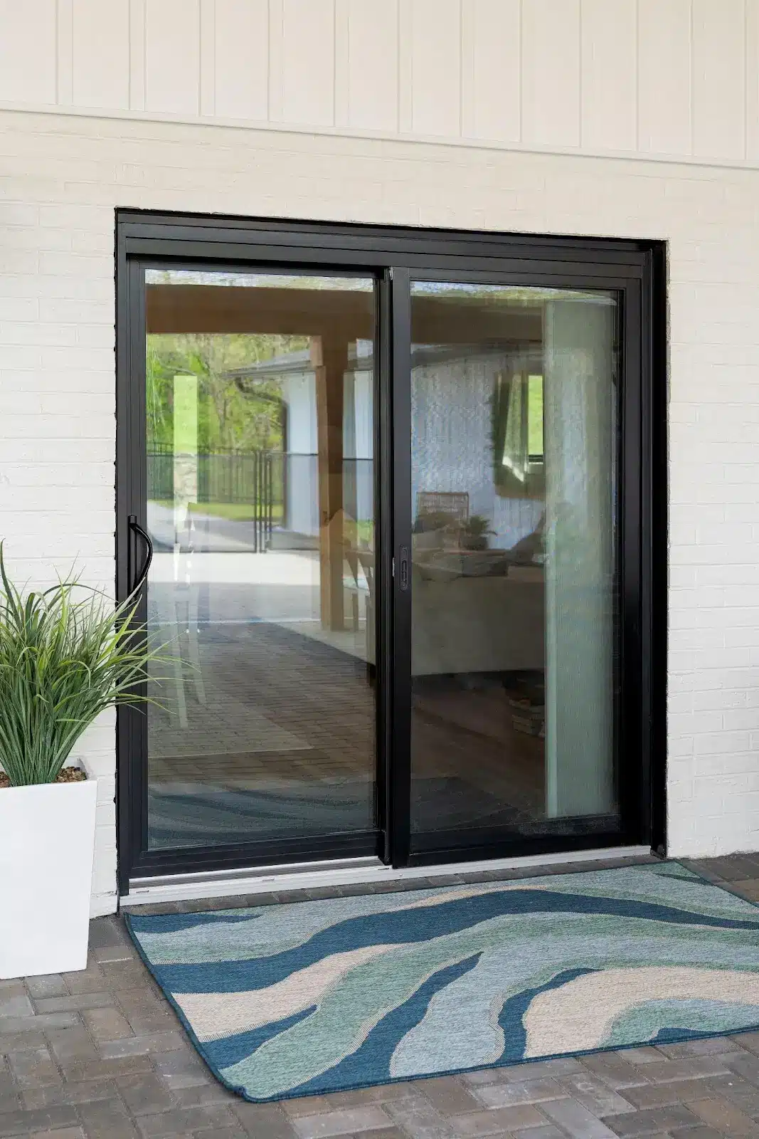 A black-framed glass sliding patio door set into a white brick wall, with a blue patterned outdoor rug and a potted plant in the foreground.
