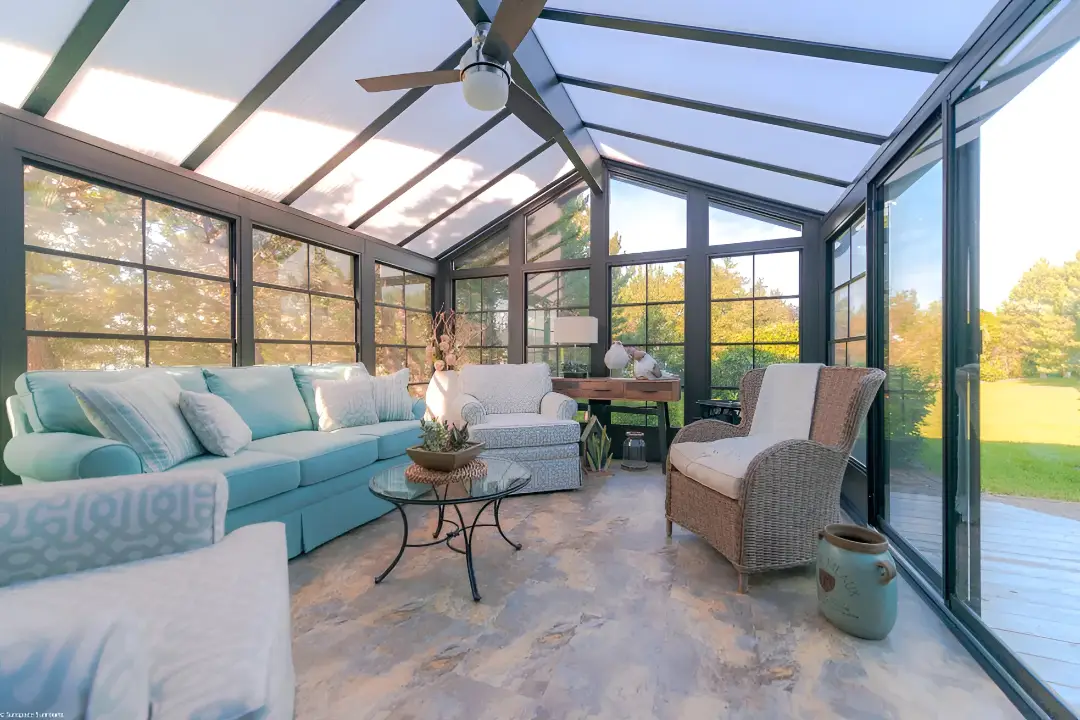 Interior of a sunroom with black vinyl frames, floor-to-ceiling windows, a vaulted translucent roof, and blue and white furniture.