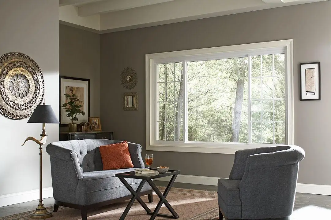 A large white sliding window in a grey living room with tufted furniture and a view of trees.