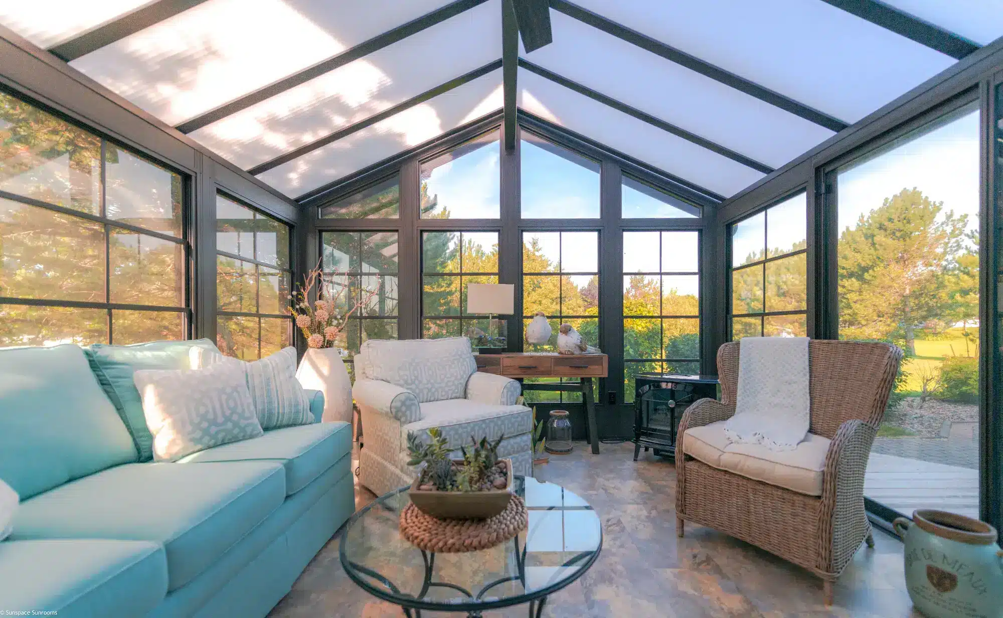 Interior of a Model 200 sunroom featuring dark bronze-colored vinyl window frames, a vaulted translucent roof, and a turquoise sofa overlooking a wooded yard.